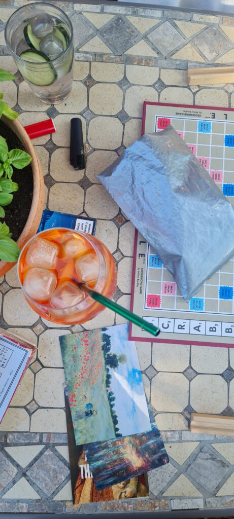 A top-down view of a table with a drink, a plant, a lighter, some postcards, and a Scrabble board, with a silver bag for the letters on top of it.
